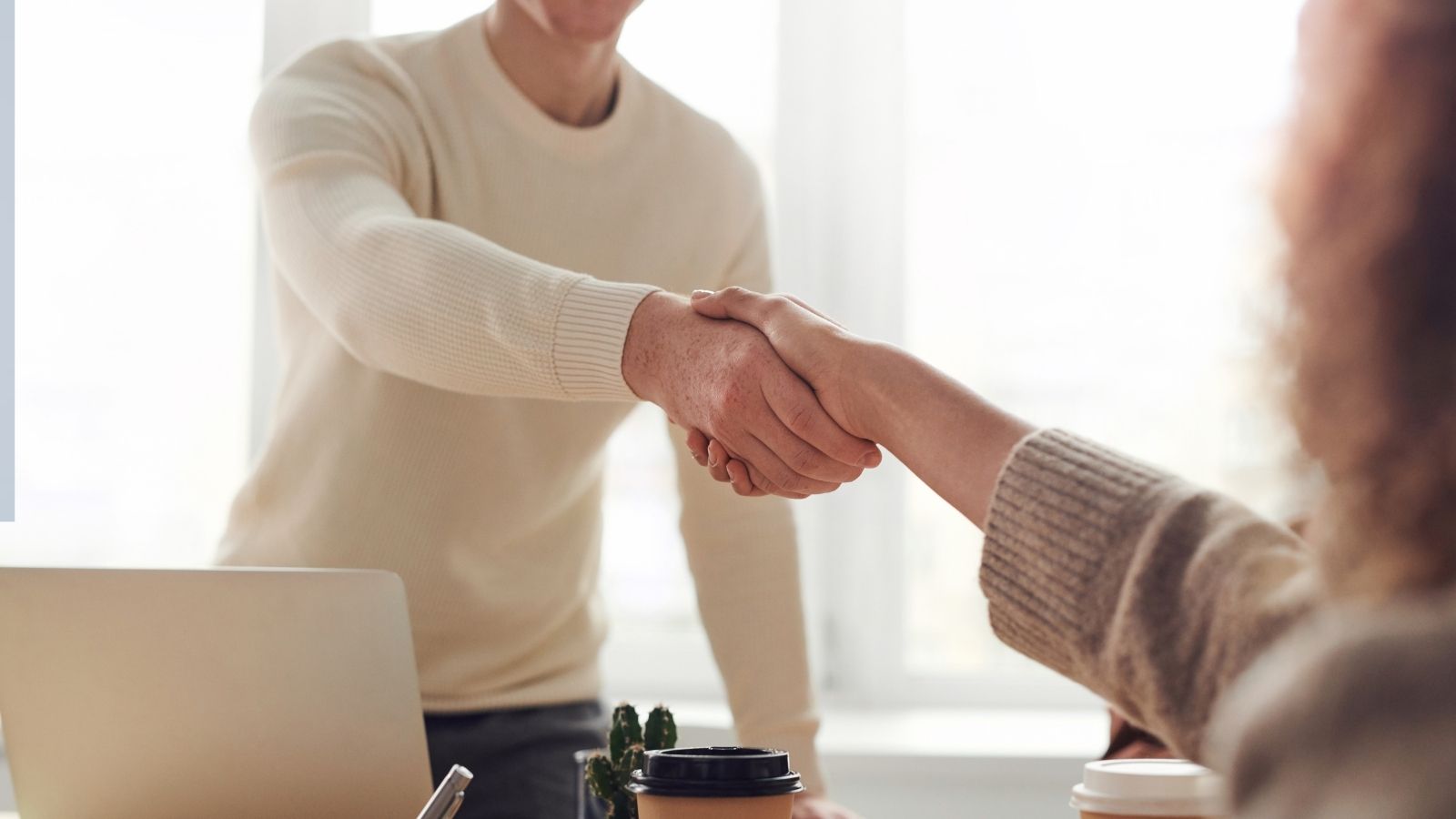 Two people shaking hands across a desk, one sitting and the other standing, with coffee cups and a laptop in view.