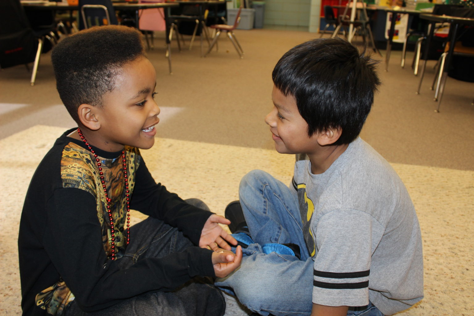 Two young boys are sitting cross-legged on the floor, smiling and talking to each other in a classroom.