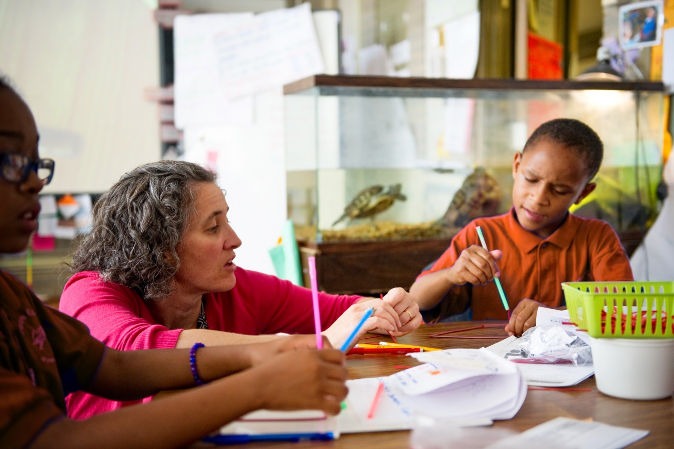 A woman assists two children with their schoolwork at a table in a classroom with a turtle tank in the background.