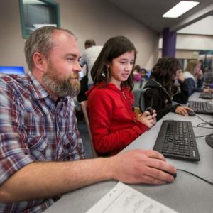 A bearded man in a plaid shirt helps a young girl in a red hoodie use a computer in a classroom setting.