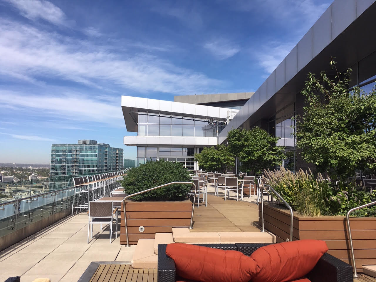 Rooftop terrace with modern seating, lush green plants, and a city skyline under a clear blue sky.