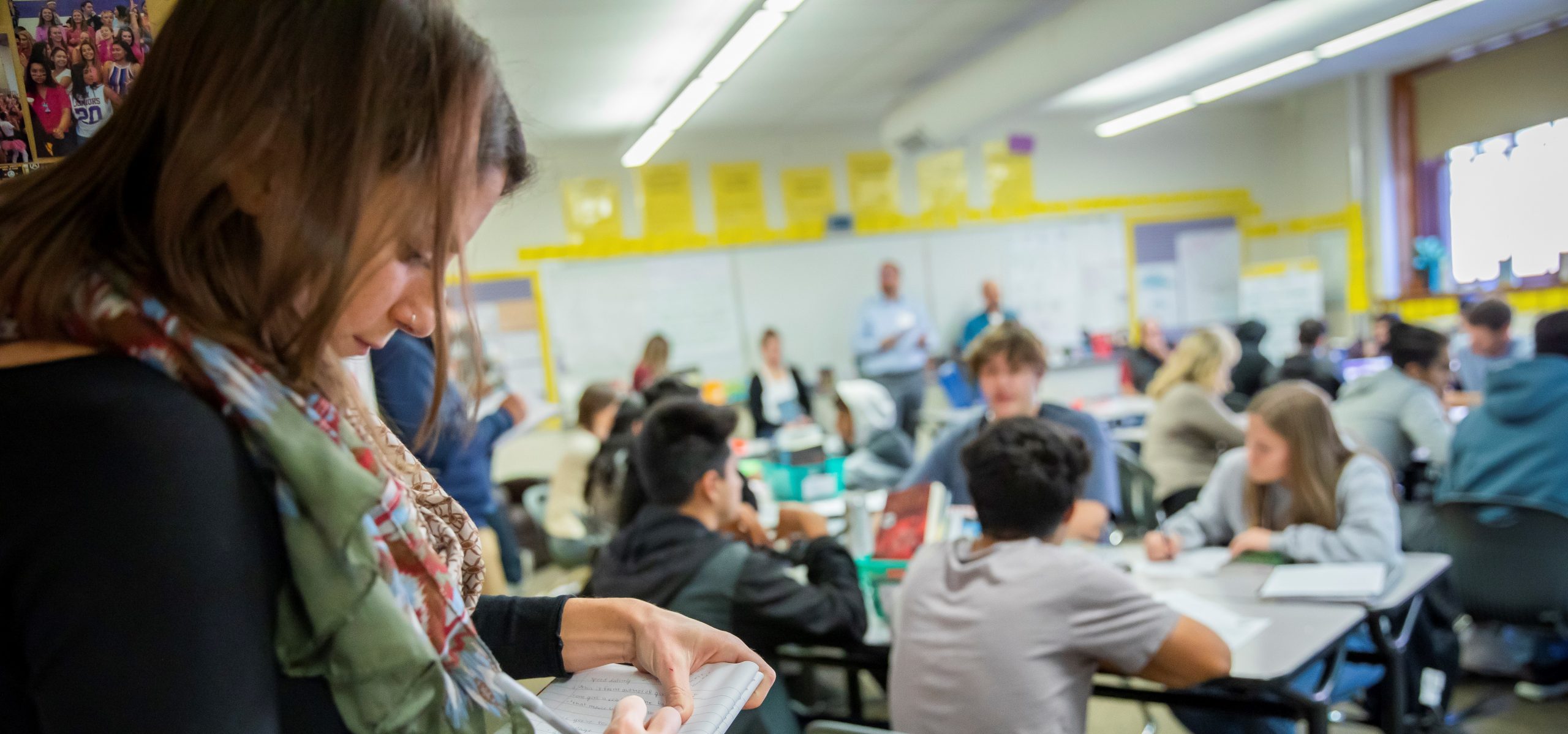 A woman writes in a notebook while students engage in group activities in a brightly lit classroom.