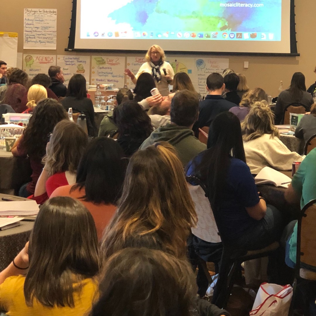 A woman speaks at the front of a classroom-style workshop with participants seated and taking notes.