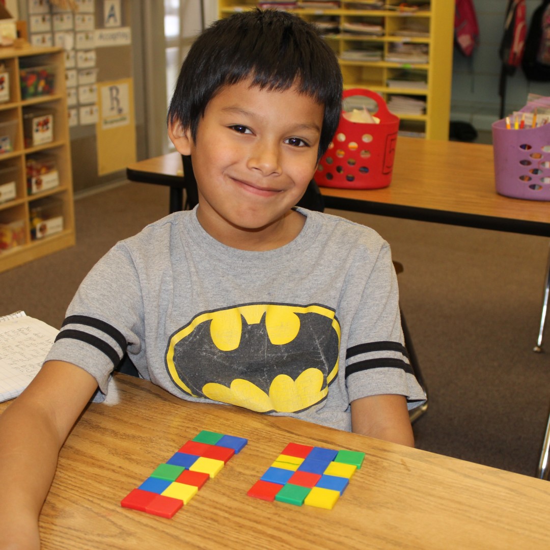 Child sitting at a desk, smiling, with colorful blocks arranged in patterns in front of them. Classroom setting.