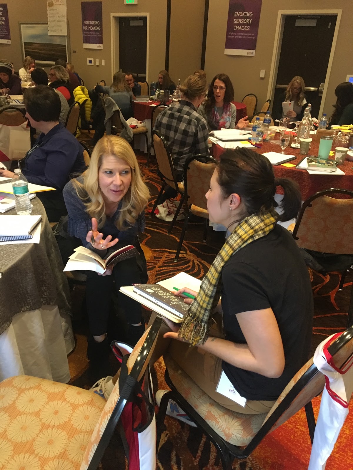 Two women sitting and discussing notes in a conference room filled with people engaged in group activities.