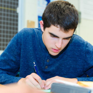 Young man in a blue shirt writing at a desk in a classroom setting.