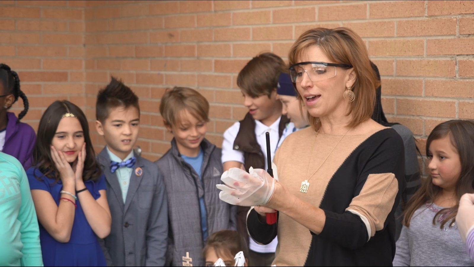 A woman wearing safety goggles and gloves demonstrates an experiment to a group of children standing against a brick wall.