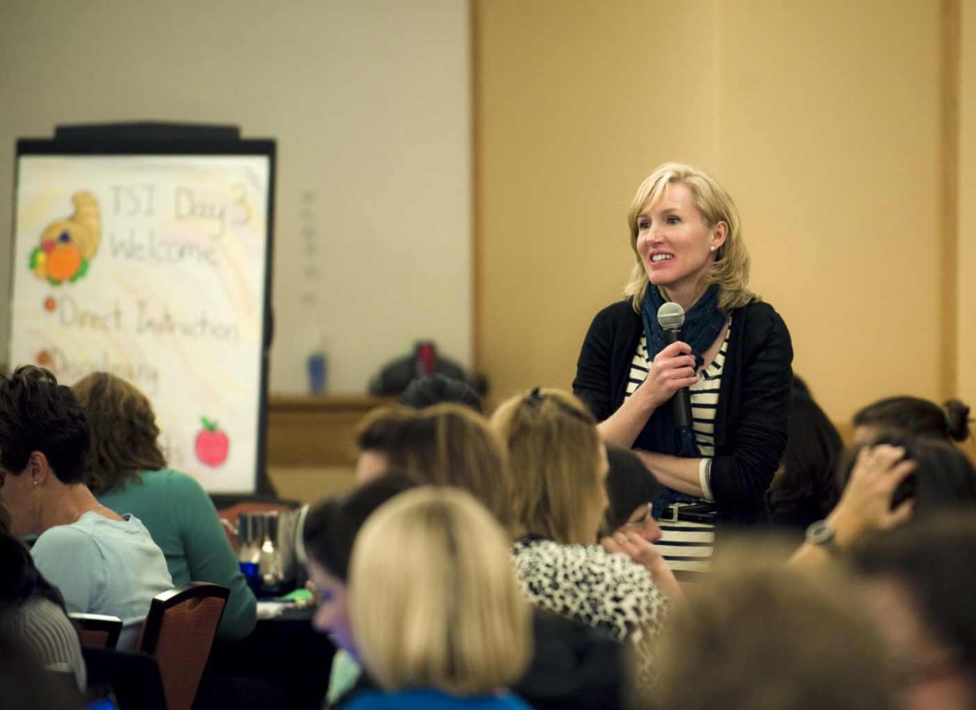 A woman with a microphone speaks to a seated audience in a room; a whiteboard with writing is visible in the background.