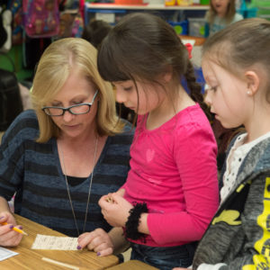 A teacher sits at a table with two young girls, helping them with a worksheet in a classroom setting.