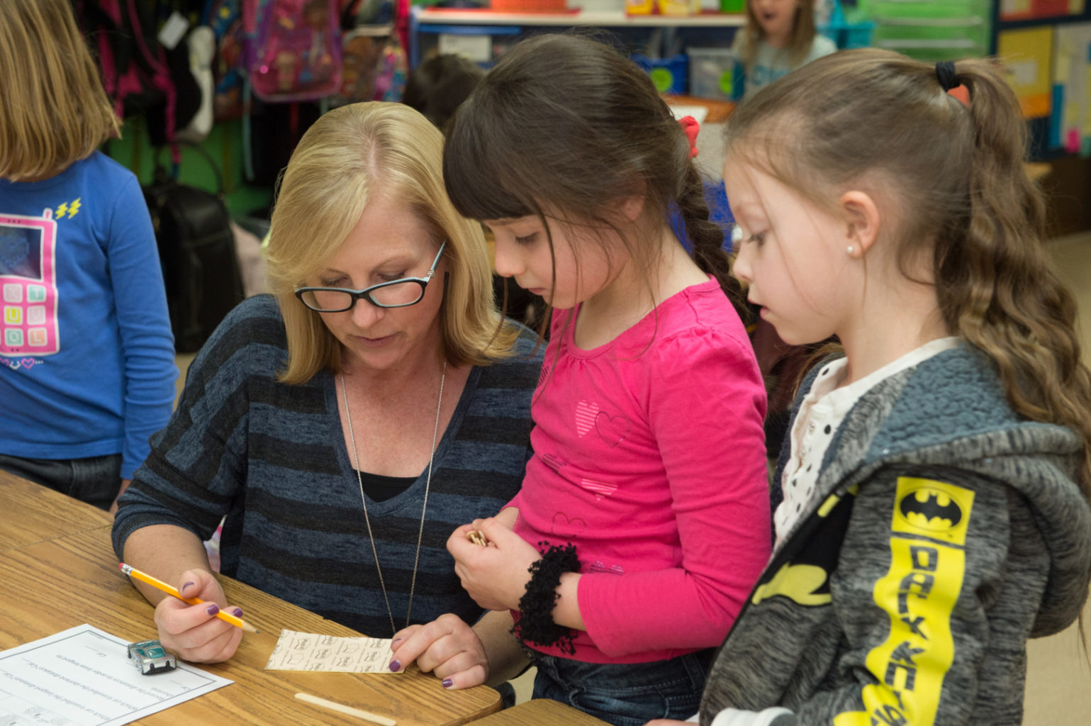 A teacher sits at a table with two young girls, helping them with a worksheet in a classroom setting.