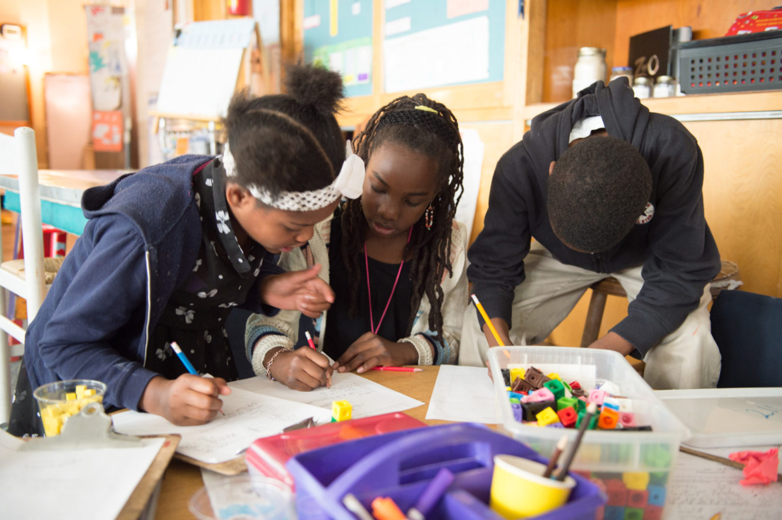 Three children are drawing and writing at a table in a classroom, surrounded by colorful papers and craft supplies.