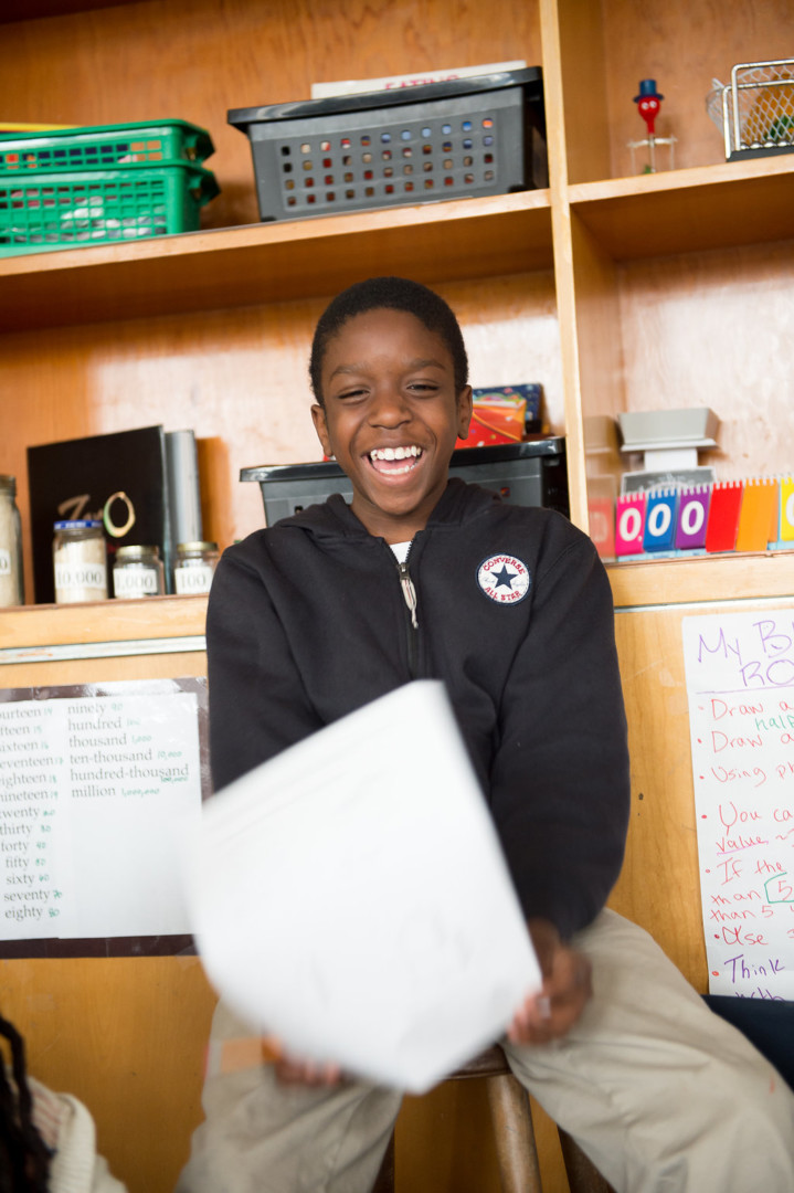 A smiling child sits in front of shelves and holds a paper, wearing a black hoodie in a classroom.