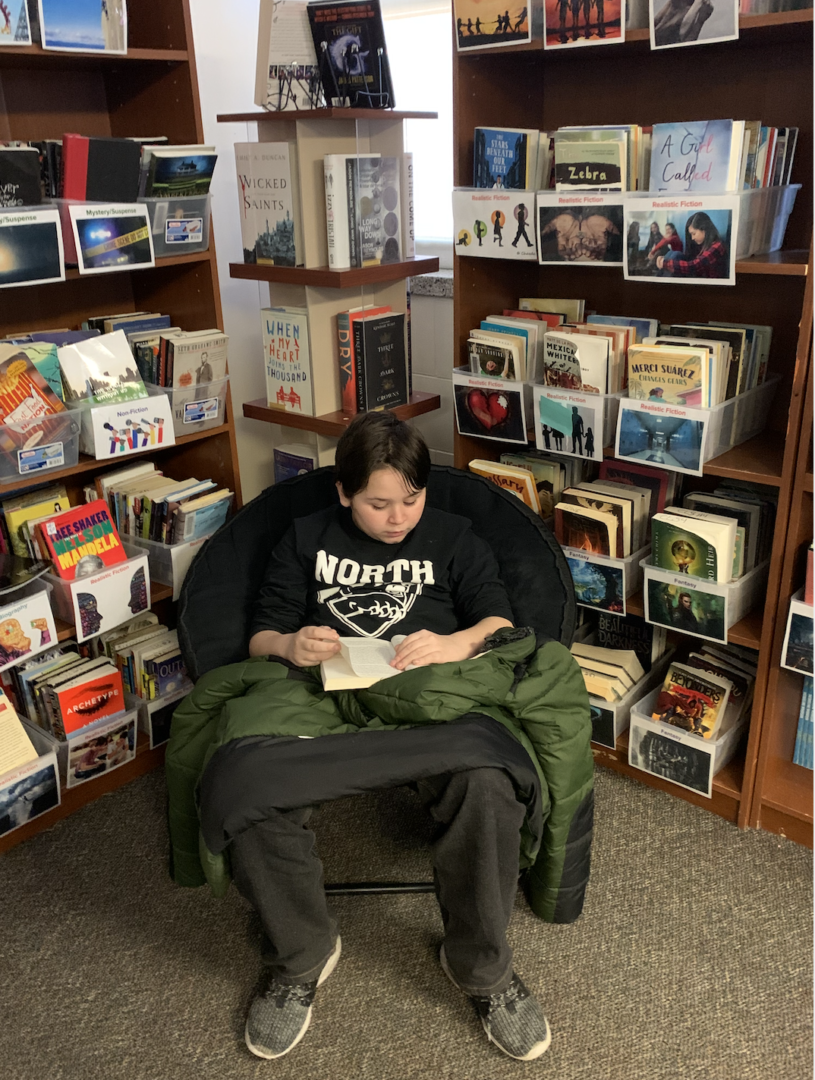 A person sits in a cozy chair reading a book, surrounded by shelves filled with books and CDs in a library or bookstore.