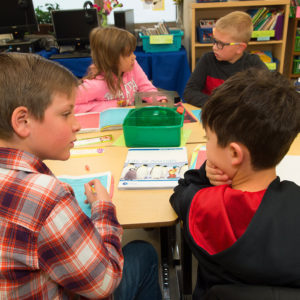 Four children sit around a table in a classroom discussing and working on a project together.