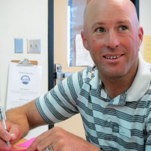 A man smiles while writing on papers at a desk, with clipboards and documents visible in the background.