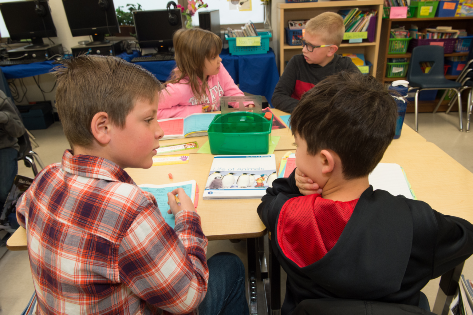 Four children sit around a table in a classroom, engaged in various activities and discussions.