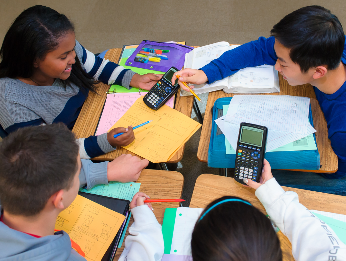 Four students collaborate at a desk with calculators, notebooks, and papers, engaged in a study session.