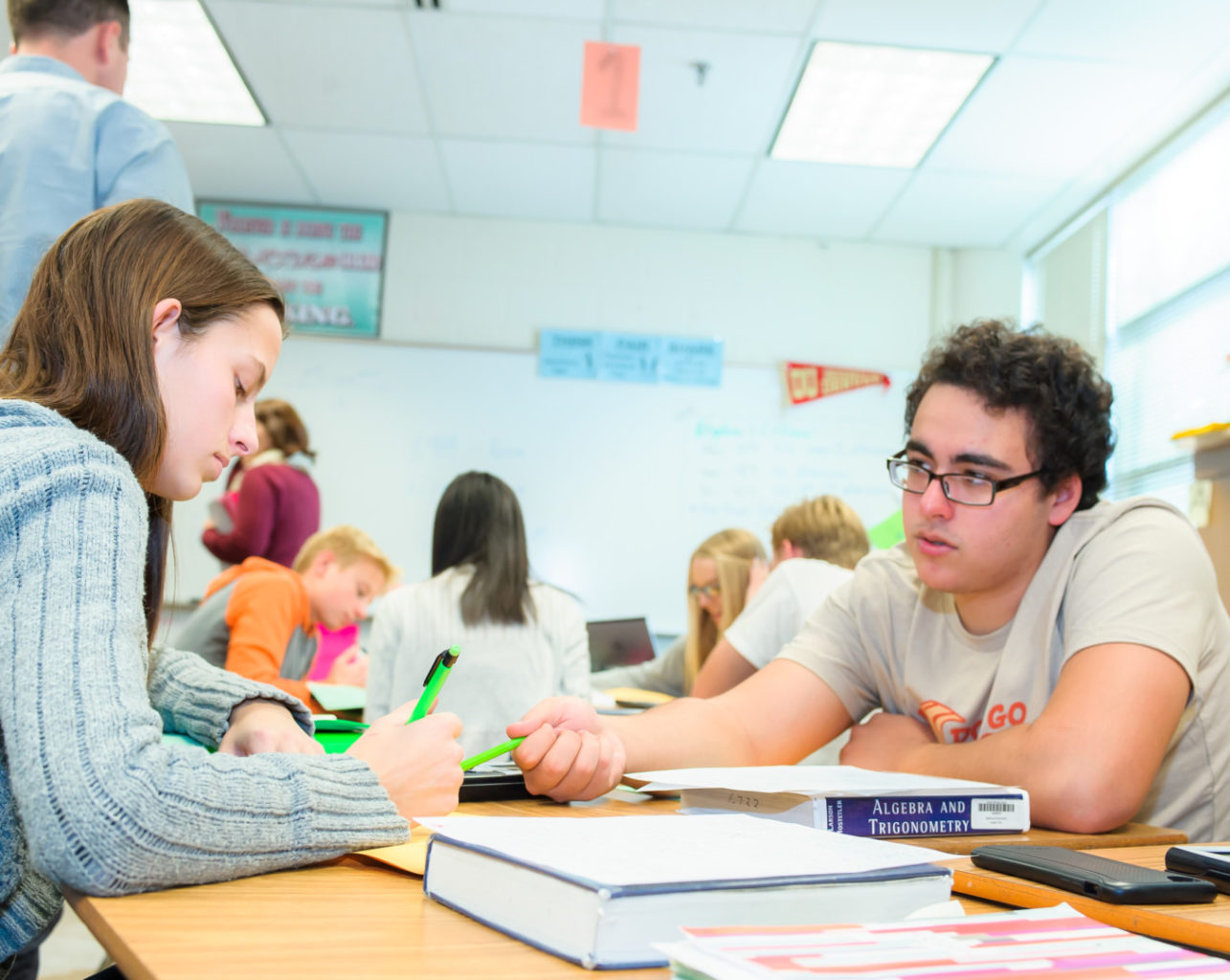 Students working together at a desk filled with textbooks and notebooks in a brightly lit classroom.