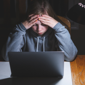 A person in a hoodie sits at a table, looking stressed with hands on their head, illuminated by a lamp, in front of a laptop.