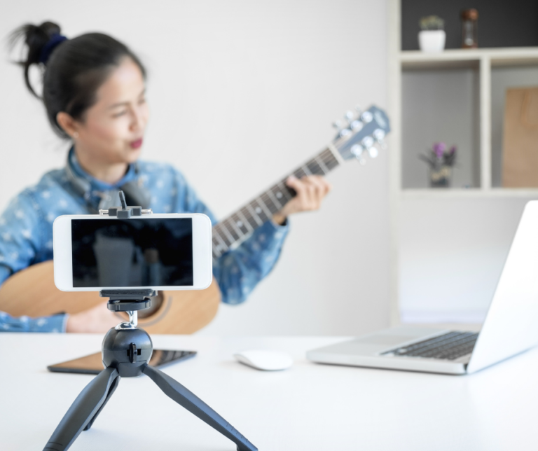 A woman playing guitar in front of a smartphone on a tripod and a laptop on a desk.