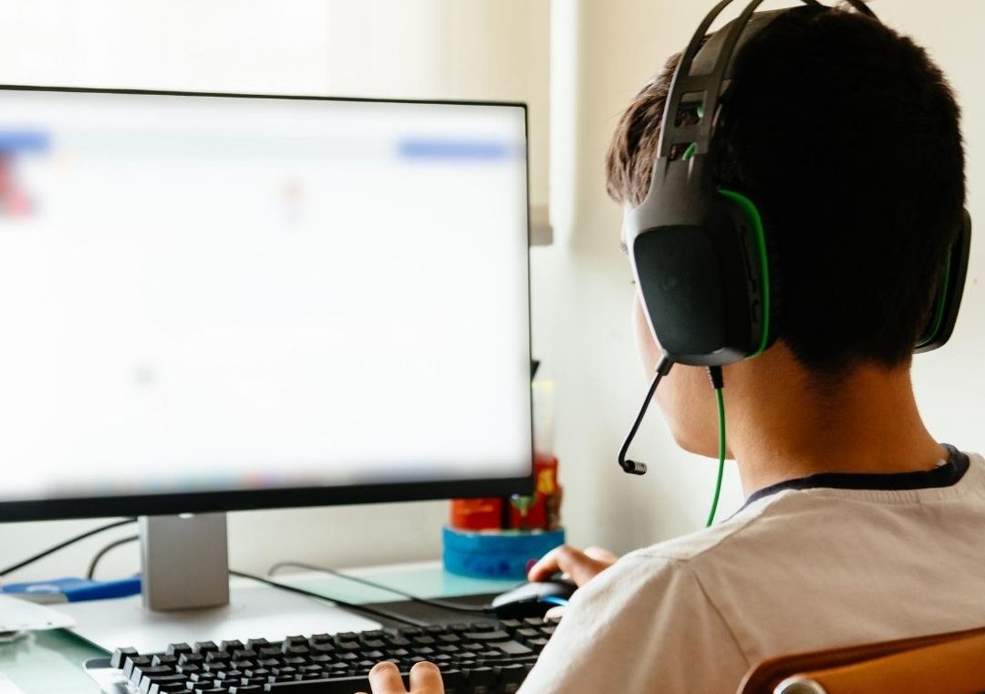 Person wearing a headset, using a desktop computer with a blurred screen, seated at a desk, focusing on the monitor.