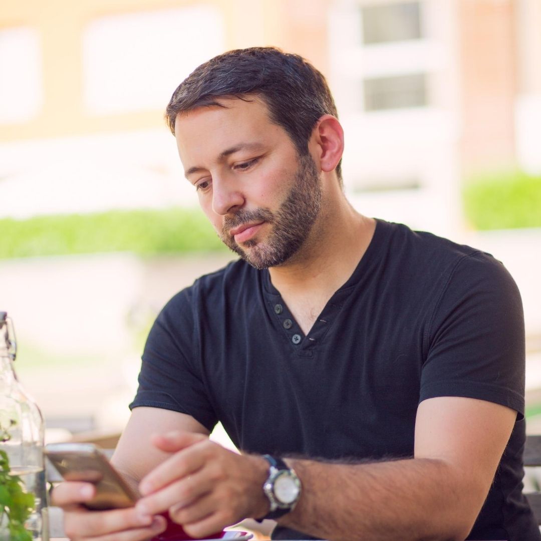 A man sitting outdoors, looking at his phone, wearing a black shirt and a watch.