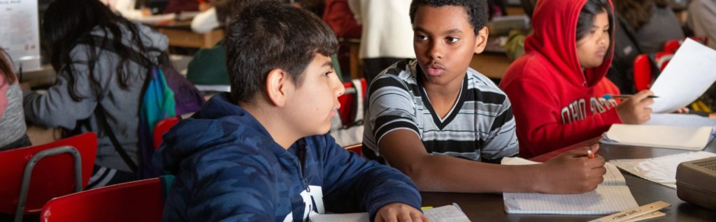 Students sitting at a table in a classroom, engaged in a discussion and working on assignments.