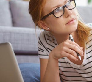 A woman with glasses and a striped shirt, holding a pen, looks thoughtful while sitting near a laptop.