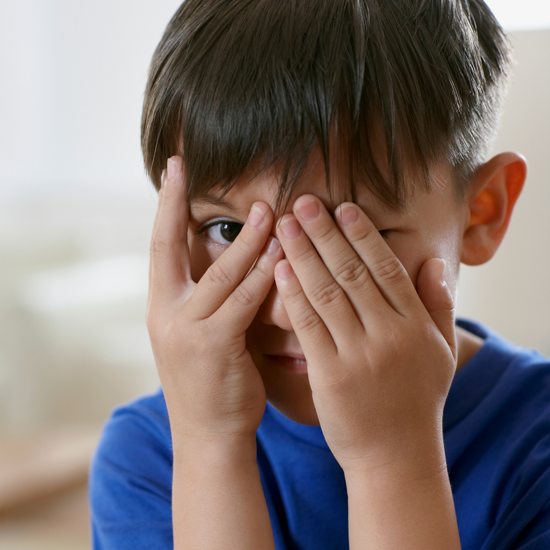 A young boy in a blue shirt peeking through his fingers as he covers his face with both hands.