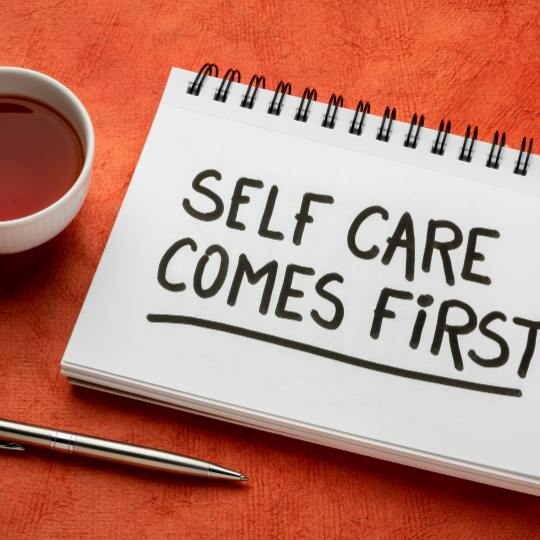 A notebook with "Self Care Comes First" written on it, a cup of tea, and a pen on a textured orange surface.