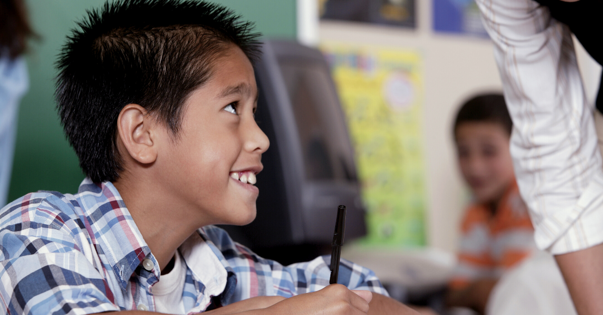 A smiling boy in a checked shirt holds a pen while looking up at an adult in a classroom setting.