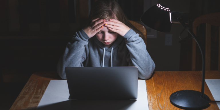 Person in a gray hoodie sits at a desk, holding their head in their hands, looking at a laptop under a desk lamp.