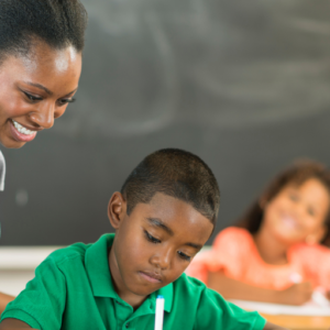 A teacher smiles while helping a student with another student working and one looking on in the background.