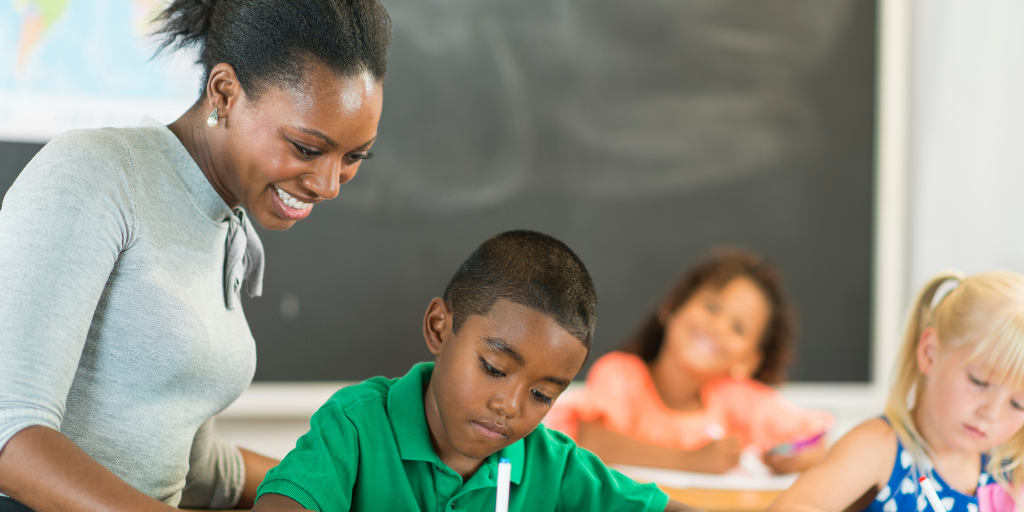 A teacher smiles while helping a student with another student working and one looking on in the background.