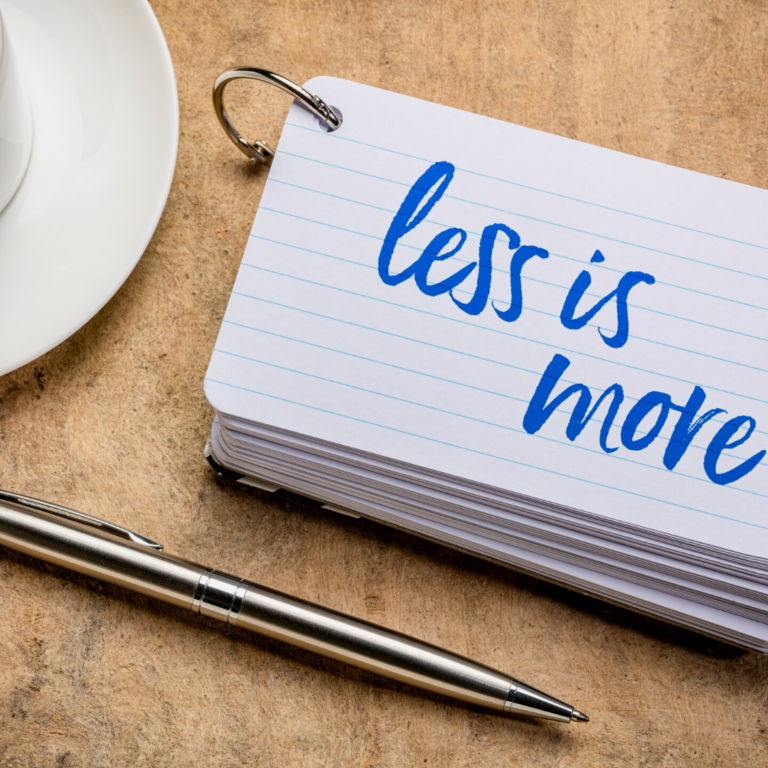 Stack of notecards on a wooden table, with "less is more" written on the top card, next to a pen and cup saucer.