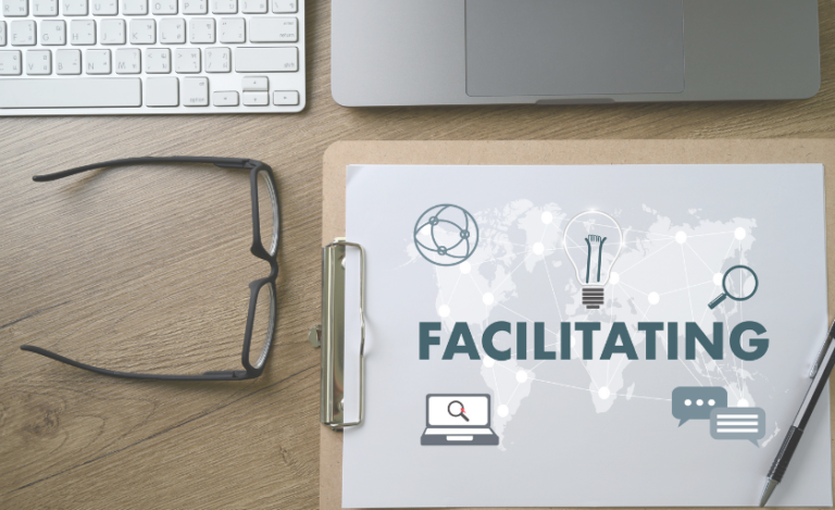 A clipboard with the word "FACILITATING" and icons, next to glasses, a pen, a keyboard, and a laptop on a desk.