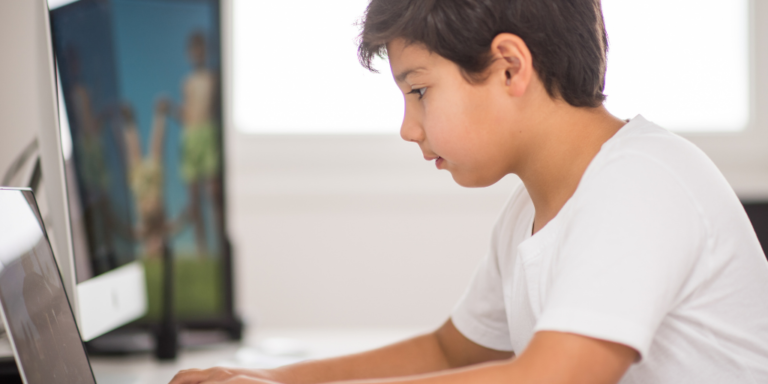 A young boy in a white shirt uses a laptop at a desk, with a blurred screen in the background.