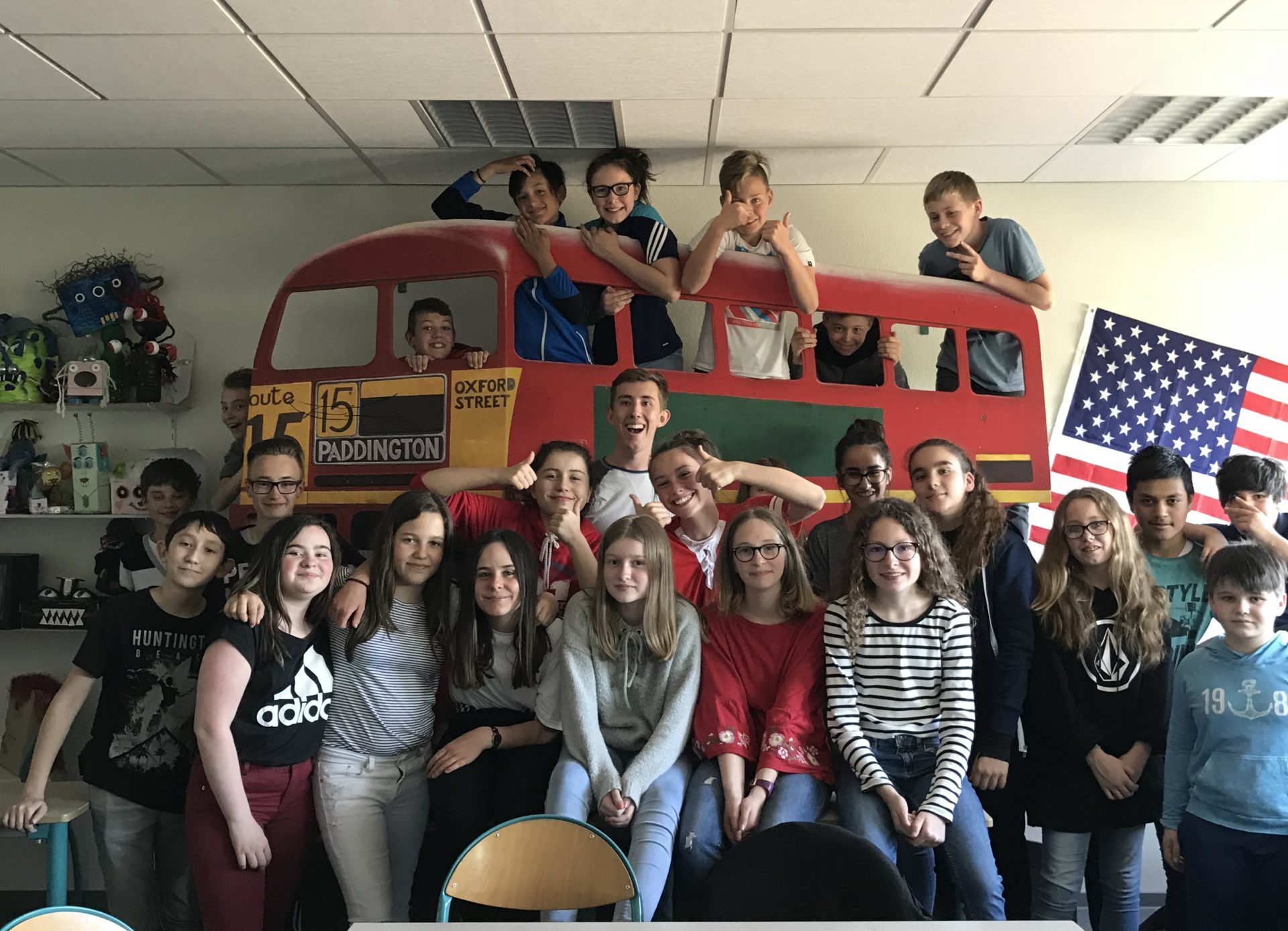 A group of students poses in front of a bus display and American flag inside a classroom.