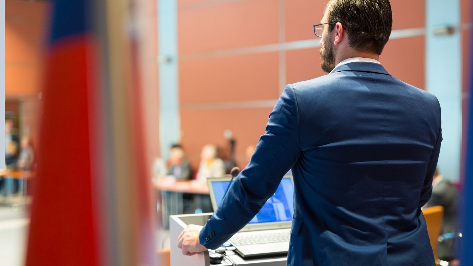 A man in a blue suit speaks at a podium with a laptop, addressing an audience in a blurred background.