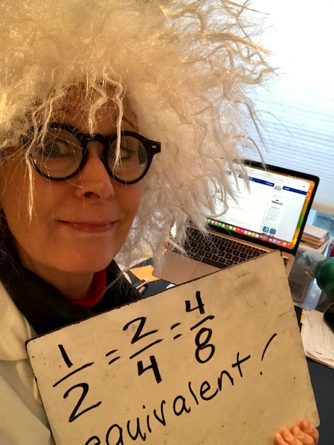 Person with wild white hair wig and glasses holds a whiteboard showing math fractions, standing near a laptop.