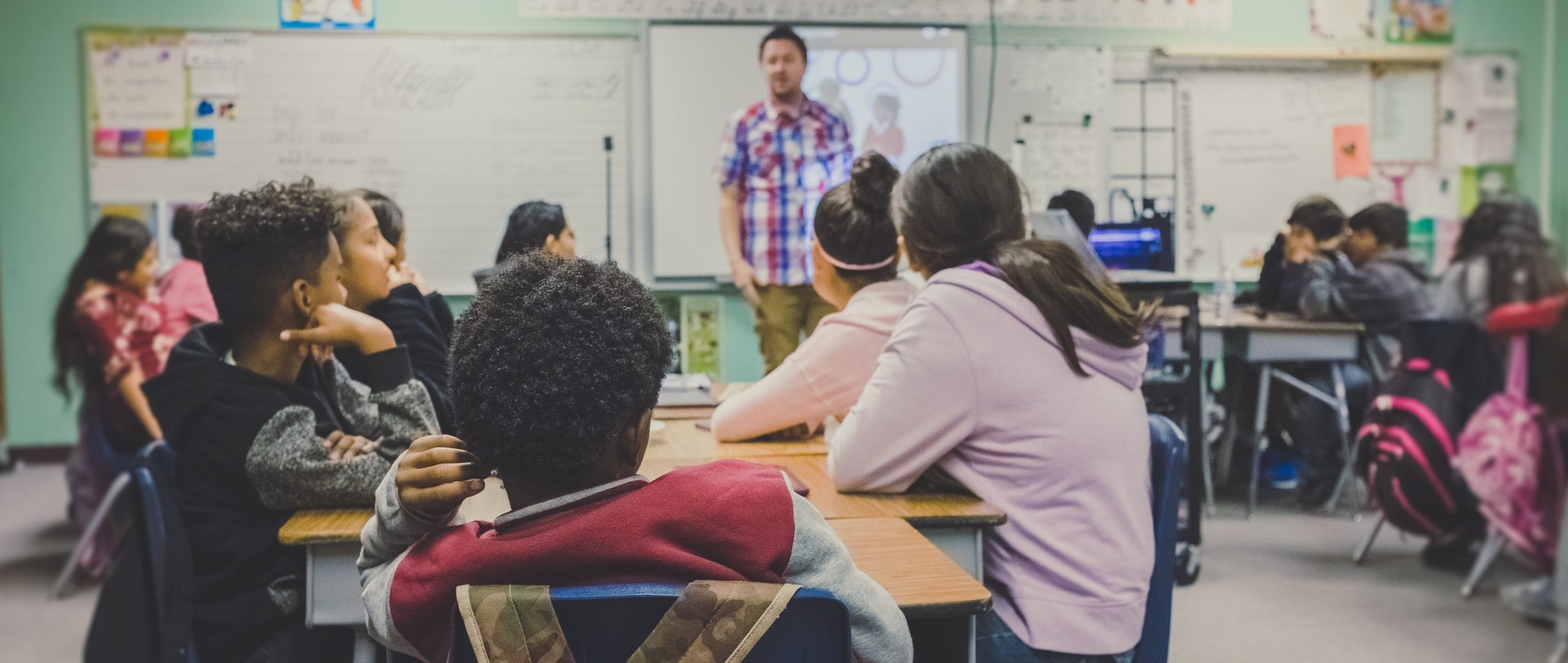 Teacher at the front of a classroom with students seated at desks, engaging in a lesson and looking attentive.