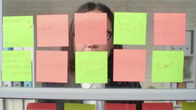 Person wearing glasses looking at a glass panel covered with colorful sticky notes in an office setting.