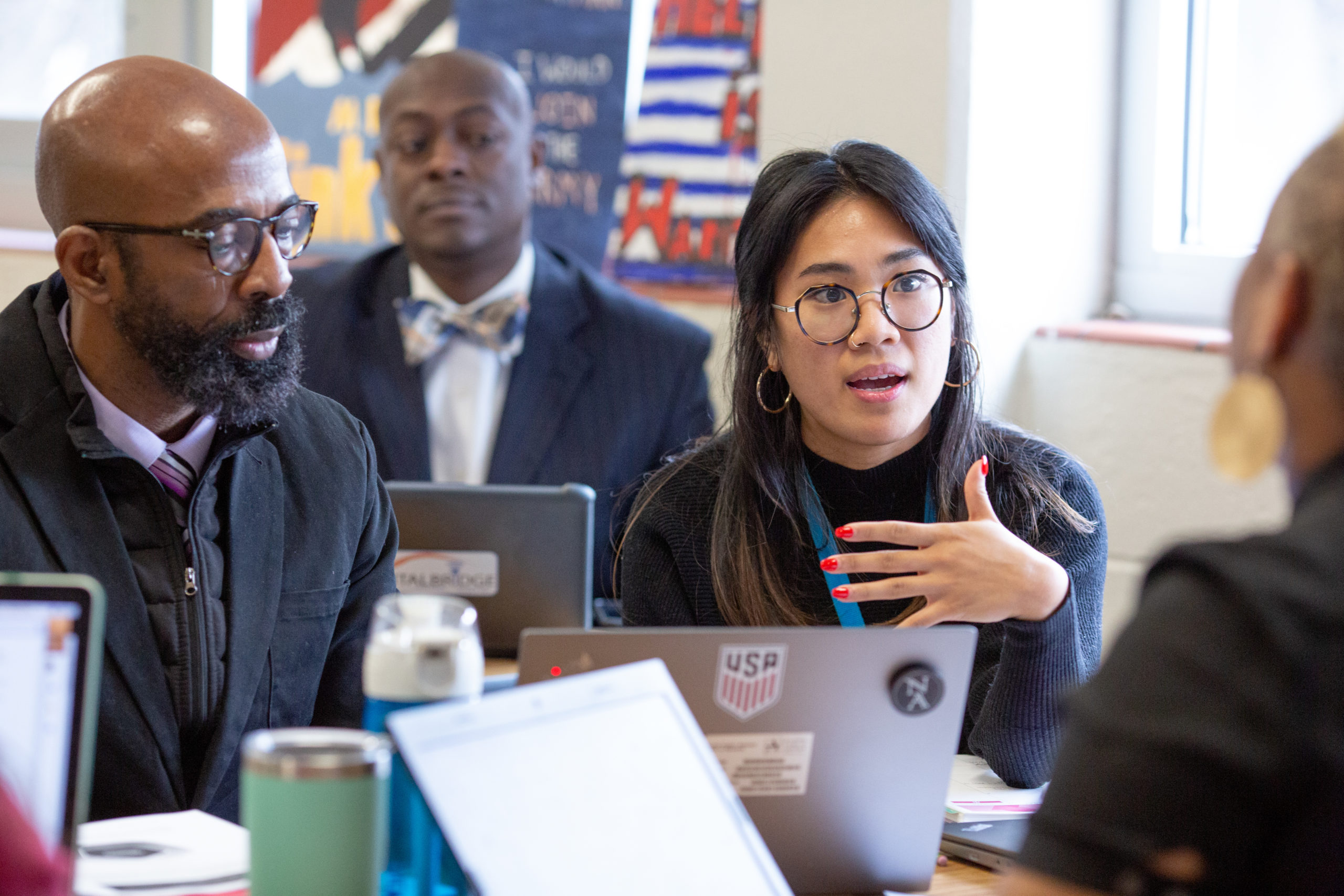 Four people in discussion around a table with laptops; one woman gestures while speaking, and others listen attentively.