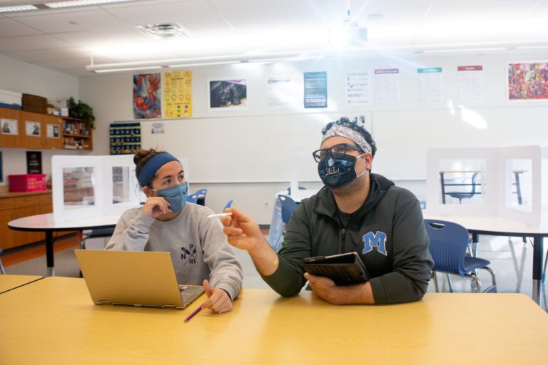 Two people wearing masks sit at a table with a laptop and tablet in a classroom, discussing something.