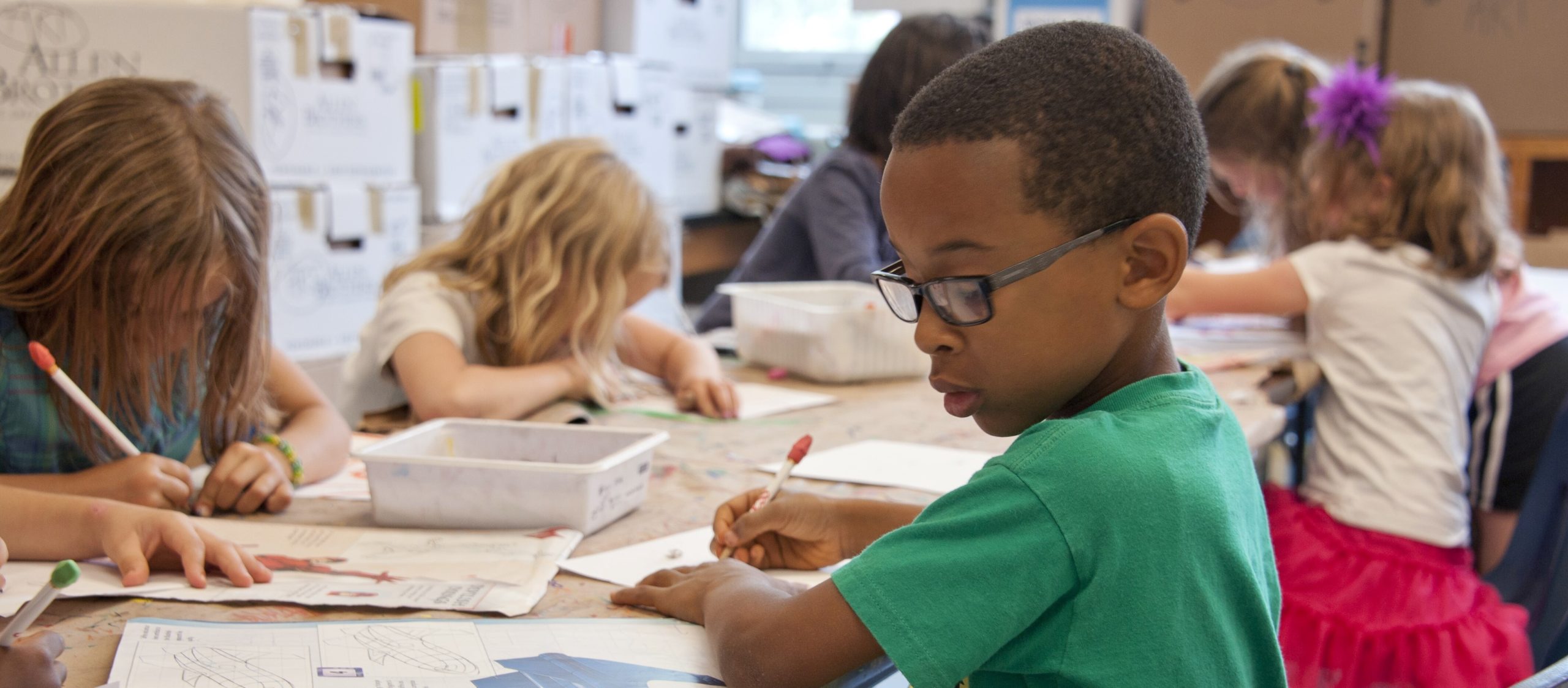 Children are seated at a classroom table, focused on writing and drawing activities.