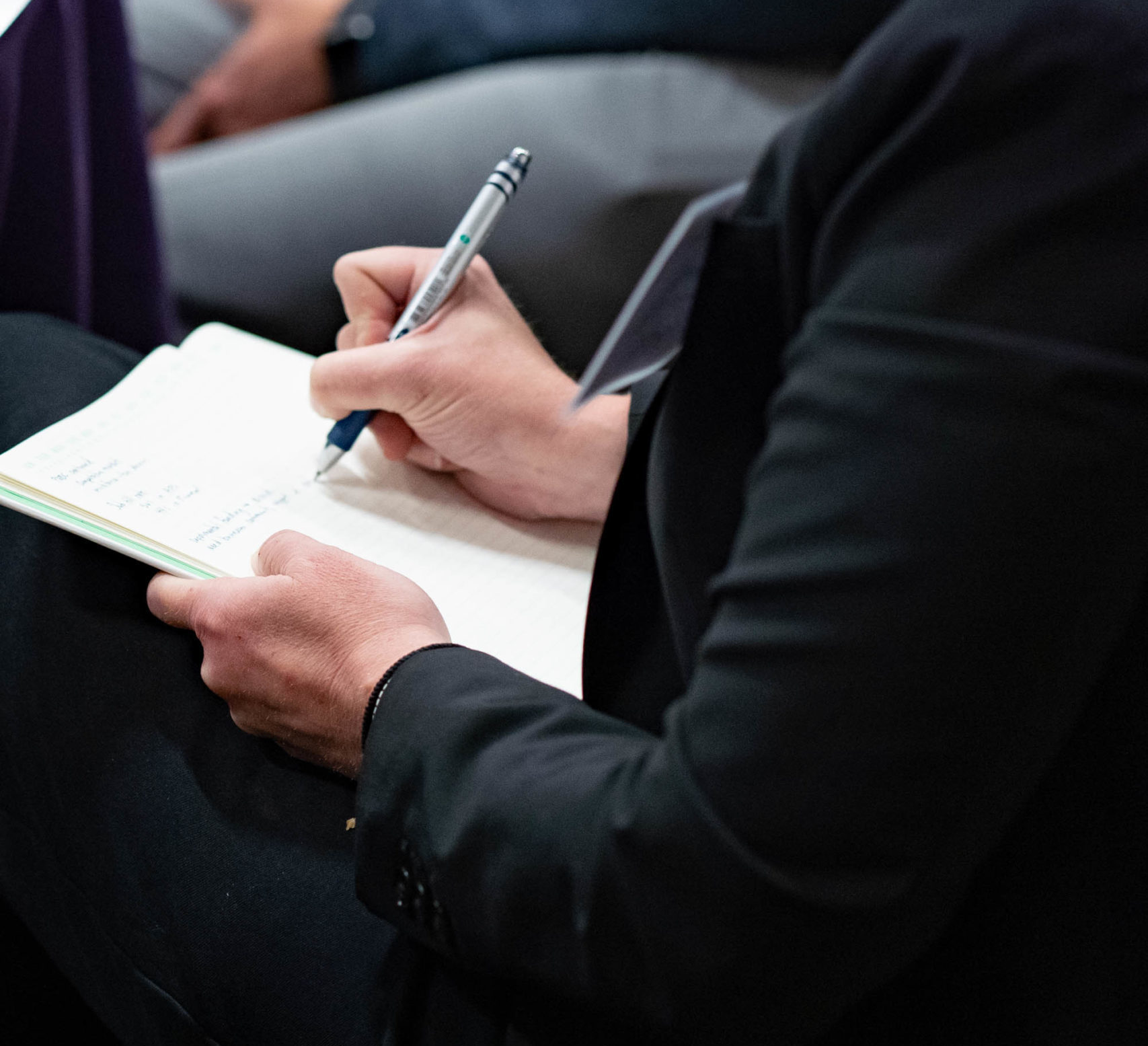 Person in a black suit jacket writing notes on a notepad with a pen.