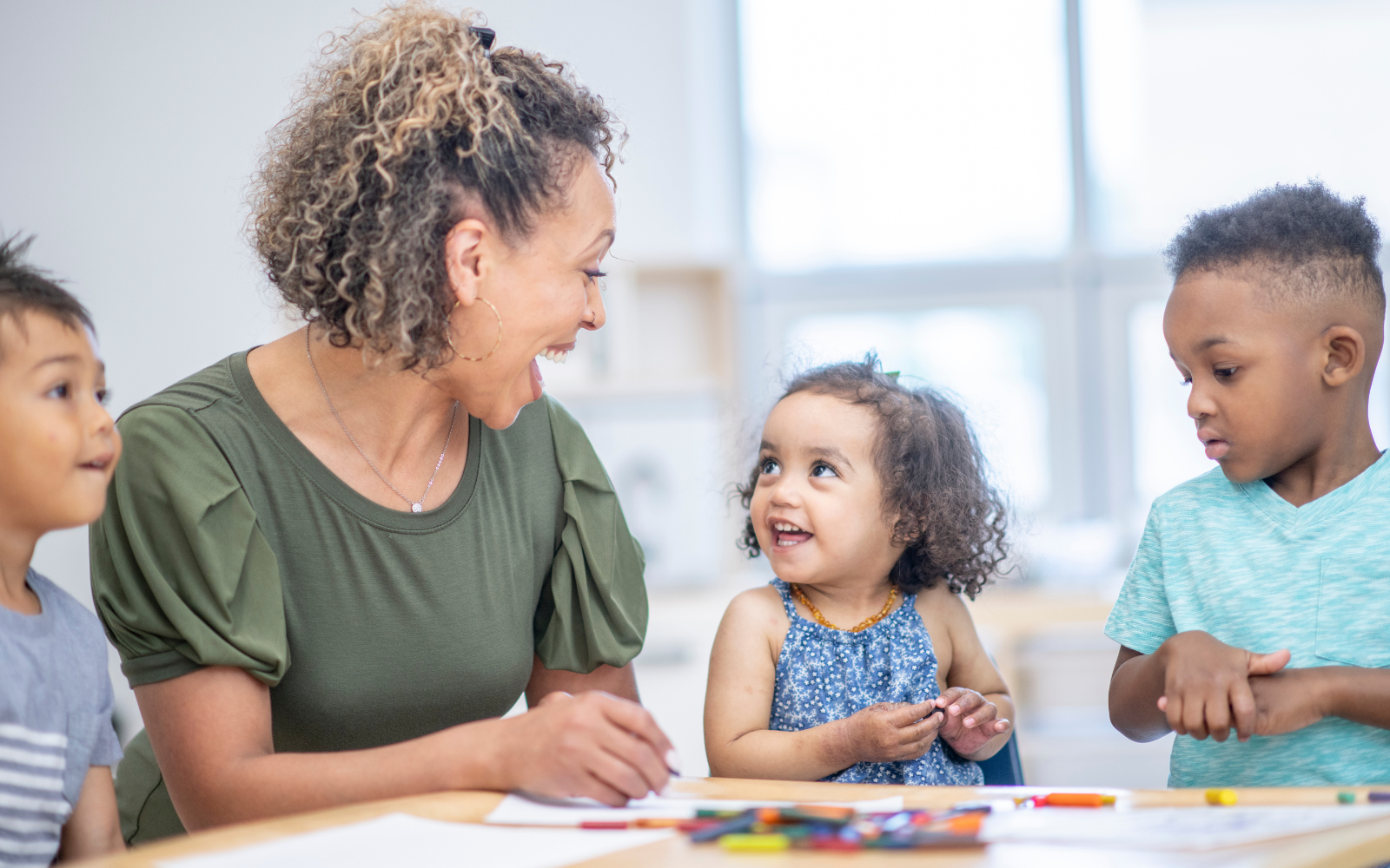 A woman engaging in a cheerful conversation with three young children in a brightly lit room with art supplies.