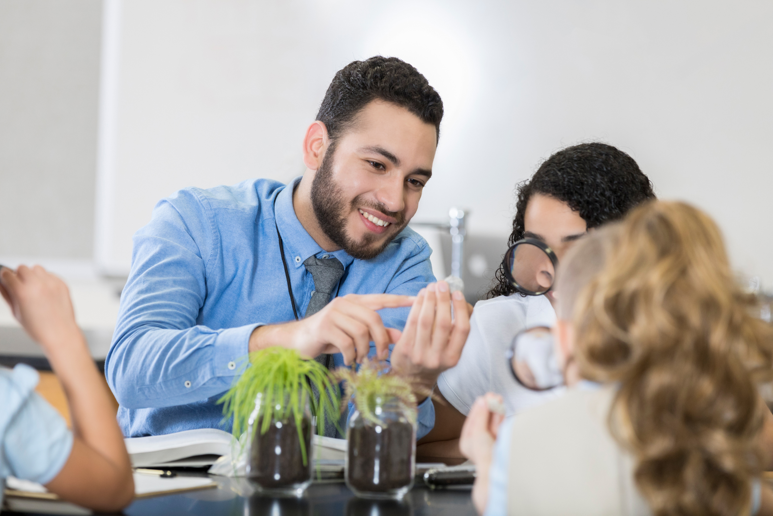 A teacher shows a plant to students in a classroom. Two jars with sprouting plants are on the table.