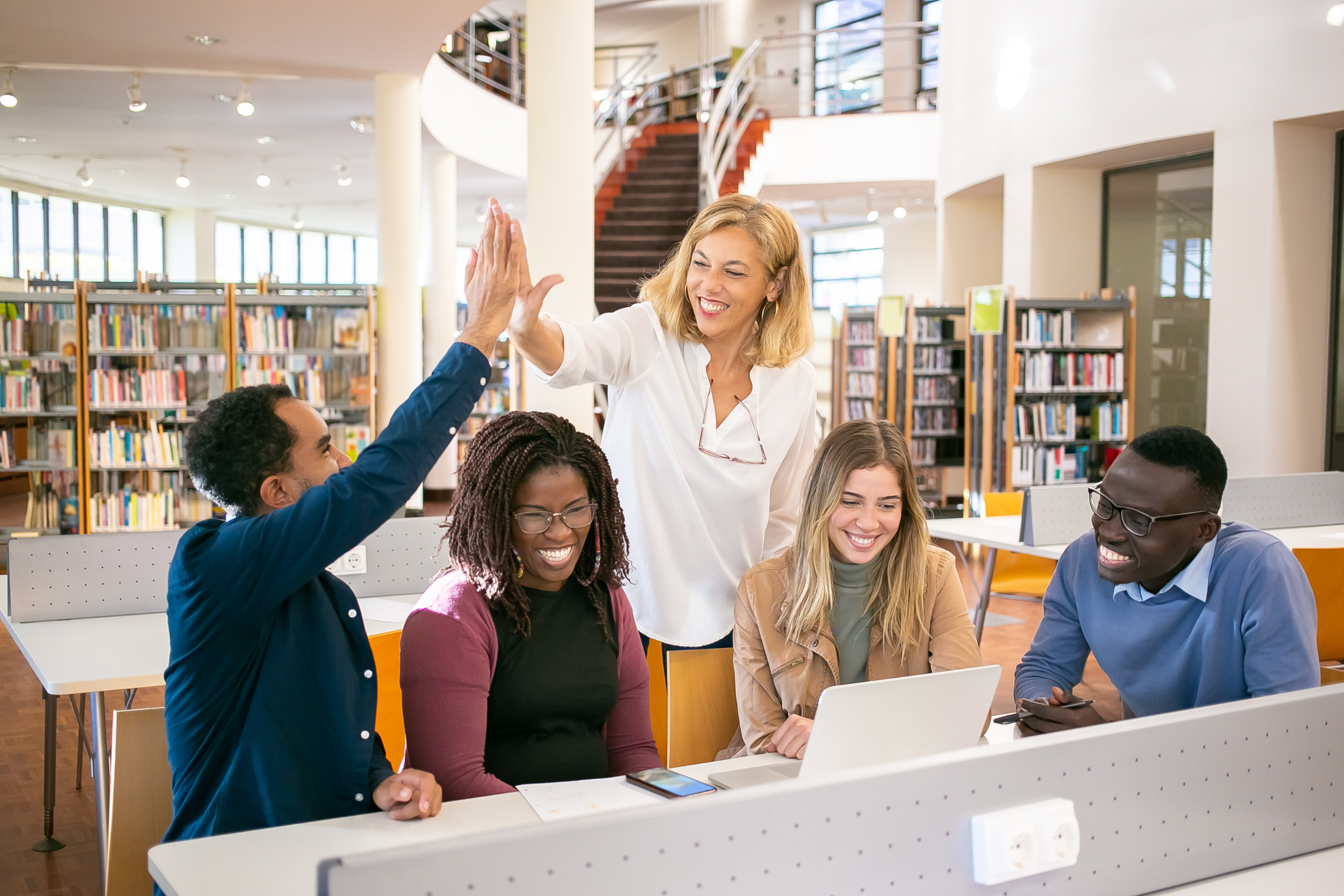 Group of five people in a library, smiling and high-fiving around a laptop, with bookshelves in the background.