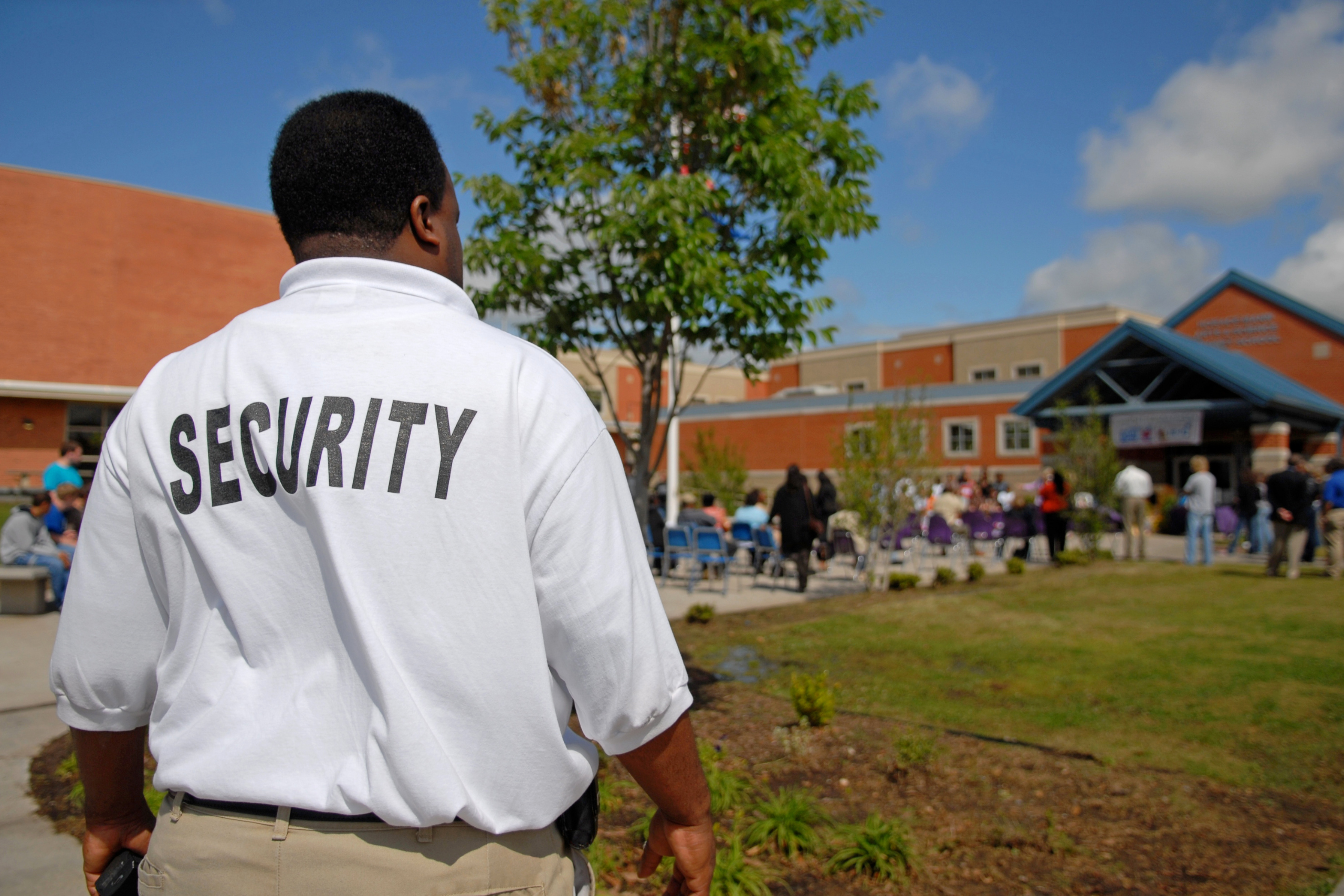 A security guard in a white shirt monitors a school event with a crowd gathered near a building on a sunny day.